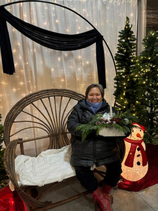 A person sitting on a bench with her centrepiece.  She is surrounded by a decorative snowman and decorated Christmas trees in the background