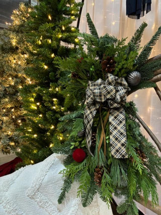 Decorative door swag with fresh mixed evergreens, a plaid bow, and pinecones, with a lit Christmas tree in the background.