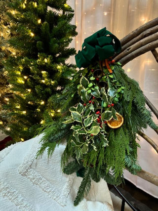 Decorative door sway made of fresh evergreens with a felt green bow and bells, dried orange slices, with a Christmas tree in the background.