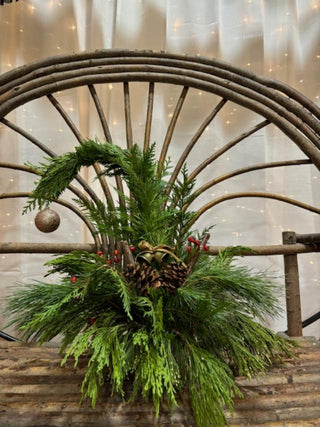 Decorative tree made with fresh greenery and pinecones on a wooden surface with a rustic wheel in the background.
